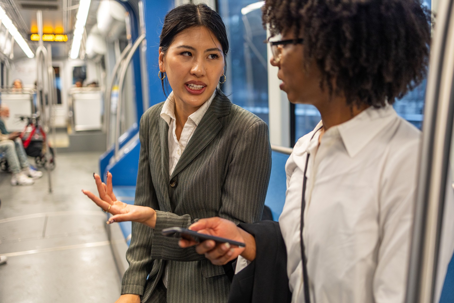 Two businesswomen, with Asian and African American ethnicity, engaged in a conversation while commuting on a city train. They appear to be discussing work-related topics in a focused and professional manner.
